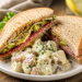 Sandwich with salami, lettuce and alpha sprouts laying on a white plate. Potato salad as a side.